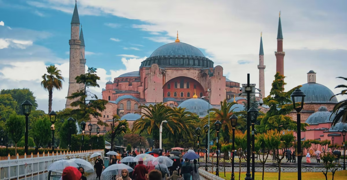 View of Hagia Sophia in Istanbul from a garden walkway on a rainy day with palm trees, street lamps, and people carrying umbrellas in the foreground.